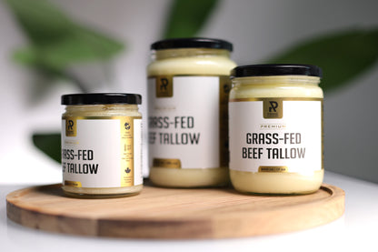 Three jars of Prime Renders grass-fed beef tallow on a wooden board with a blurred green leafy background.